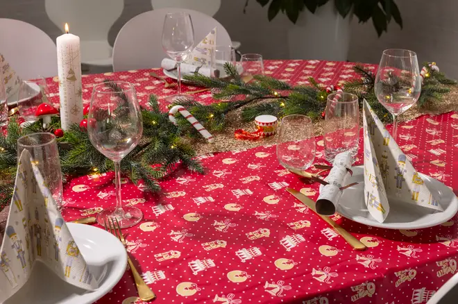 Rødt juledug med hvide og gyldne festlige mønstre, dækket på spisebord med tallerkener, guldbestik, foldede servietter, vinglas og gran-guirlande som midterpynt.