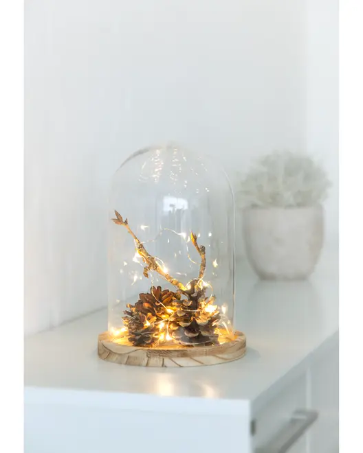 Decorative arrangement featuring pinecones and twigs placed on a round wooden base, enclosed in a clear glass dome. The pinecones and twigs are wrapped with warm white LED fairy lights, creating a cozy and inviting glow. The overall design is rustic and elegant, suitable for home decor or as a centerpiece.