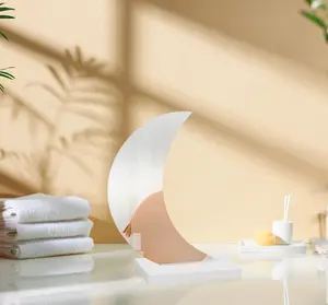 Dekorativt spegel formad som en halvmåne med vit bas, placerad på badrumsbänk. Omgiven av vikta handdukar och toalettartiklar, perfekt för modern inredning.