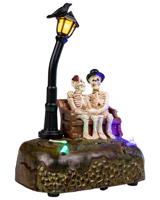 Halloweenfigur med två skelett på bänk under sned gatlykta, färgglada LED-lampor på texturerad bas, iförda hattar och accessoarer.
