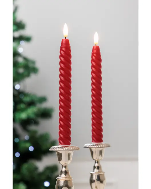 To røde spiralformede stagelys, tændte, placeret i sølvlysestager. Elegant snoet design, velegnet til festlige eller dekorative lejligheder. Sløret juletræ i baggrunden.