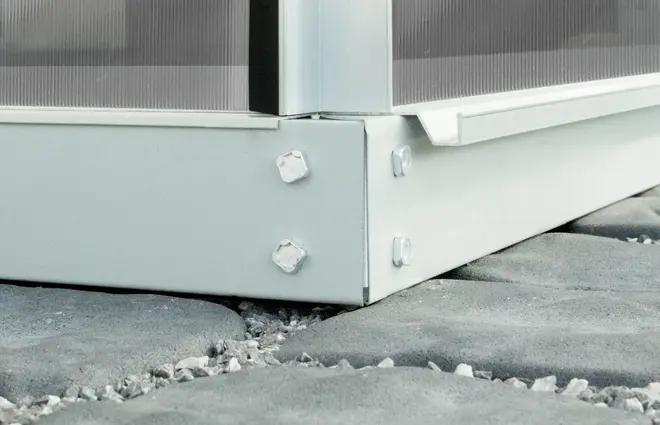 Närbild av metallram till växthus med bultar, säkert fastsatt i hörnet, placerad på stenplattor. Hållbar konstruktion för stabil växthusinstallation.