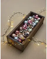 Rektangulär trälåda fylld med blandade julgranskulor i metallic rosa, guld, silver och grönt. Omgiven av varmvita ljusslingor. Kulor och kotteformade dekorationer.