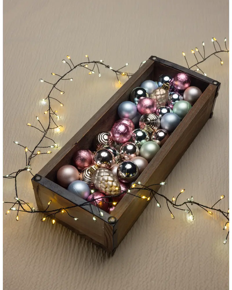 Rektangulär trälåda fylld med blandade julgranskulor i metallic rosa, guld, silver och grönt. Omgiven av varmvita ljusslingor. Kulor och kotteformade dekorationer.
