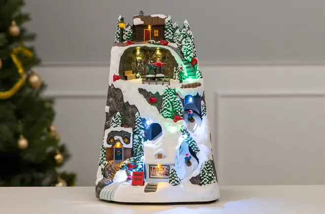 Oplyst julelandsbydekoration med snedækket bjerg, fyrretræer, hytter, skiløbere og farverige LED-lys. Detaljeret vinterscene, ideel til bord- eller pejseudsmykning i juletiden.