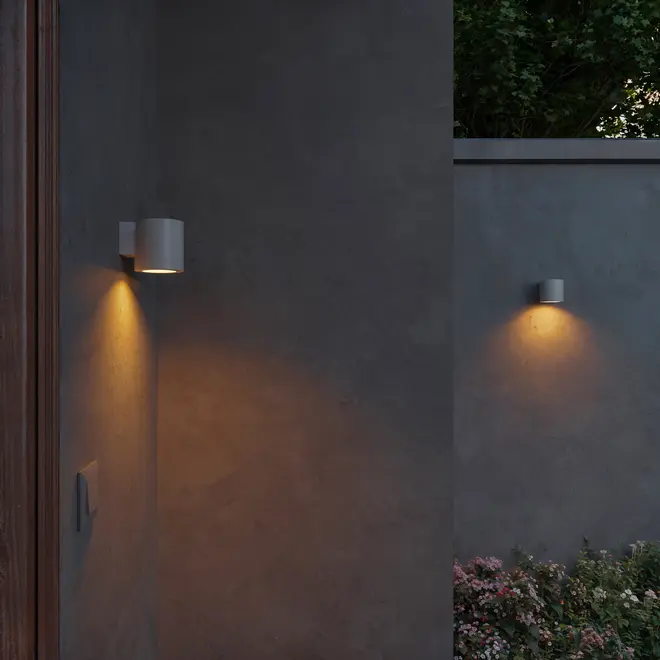 Moderne cylindriske udendørs væglamper i mat grå finish, monteret på betonvæg, udsender varmt nedadgående lys. Velegnet til have eller terrasse, vejrbestandigt design.