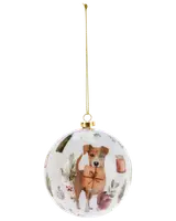Rund vit julgranskula med målad hund som håller ett paket, omgiven av grönska, ljus och juldekorationer. Har guldfärgad upphängning och snöre.