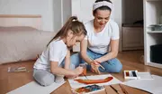 Ett barn och en vuxen sitter på golvet och målar med akvarellfärger. Konstmaterial, penslar och papper ligger runt dem. Båda har avslappnade kläder och fokuserar på sitt konstverk.