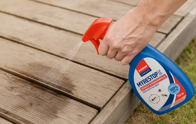 Blå sprayflaske med rød dyse, mærket "MyreStop" mod myrer, 750 ml. Anvendes udendørs, her på træterrasse, hvor væske sprayes ud.