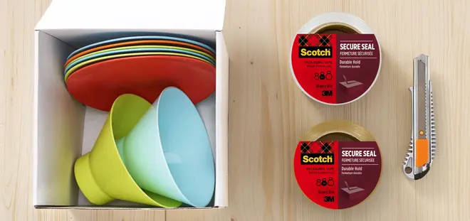 Sæt med farverige plastik tallerkener og skåle i en kasse, to ruller Scotch Secure Seal pakketape og en hobbykniv på lyst træbord.