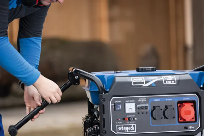 Bærbar generator med blå og sort kabinet, flere strømudtag, kontrolpanel og solidt håndtag. En person samler eller justerer håndtaget til transport eller opbevaring.