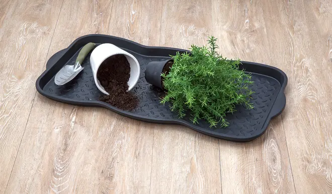 Svart plastbricka med upphöjda kanter och mönstrad yta, innehåller liten planteringsspade, utspilld jord, vit kruka, svart kruka och grön krukväxt.