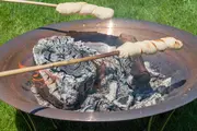 To pinde med brøddej ristes over et bålsted med brændende træ og gløder, ideelt til udendørs madlavning eller camping.