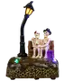 Halloweenfigur med två skelett på bänk under böjd gatlykta, färgglada LED-lampor på texturerad bas. Ena skelettet har hatt, det andra röd rosett.