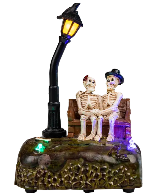 Halloweenfigur med två skelett på bänk under böjd gatlykta, färgglada LED-lampor på texturerad bas. Ena skelettet har hatt, det andra röd rosett.