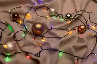 Set med guld- och silverfärgade julkulor samt en slinga med flerfärgade LED-lampor, placerade på beige tyg. Perfekt för juldekoration.