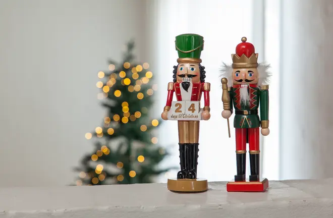 Två trädockor i form av nötknäppare, en håller en skylt med '24 dagar till jul.' Båda är målade i rött, grönt och guld, stående på vit yta.