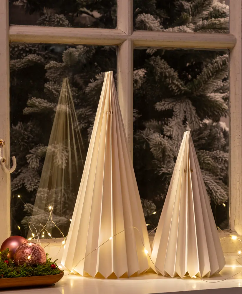 Two decorative cone-shaped ornaments made of folded white paper, resembling stylized Christmas trees. The ornaments have a pleated design and are illuminated from within, creating a warm glow. They are placed on a windowsill with string lights and festive baubles nearby, with a snowy tree visible outside the window.