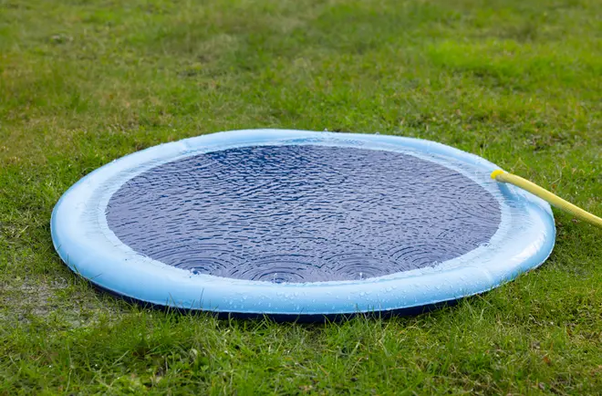 ,Rundt blåt oppusteligt soppebassin fyldt med vand, placeret på grønt græs. En gul haveslange fylder bassinet. Velegnet til små børn eller kæledyr udendørs.