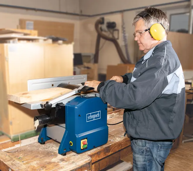 Elektrisk Scheppach høvle-tykkelseshøvl med blåt kabinet, sikkerhedsafbryder og justerbar anslag, bruges til træbearbejdning for at høvle og glatte brædder. Vist i brug på værkstedsbord.