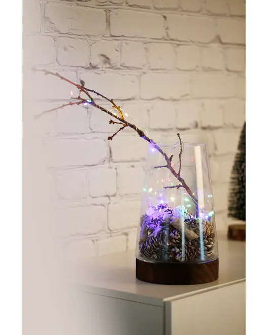 Decorative arrangement featuring a clear glass vase with a dark wooden base, filled with pinecones and a bare branch. The branch is wrapped with multicolored string lights, creating a festive and cozy ambiance. The vase is placed on a light-colored surface against a white brick wall.