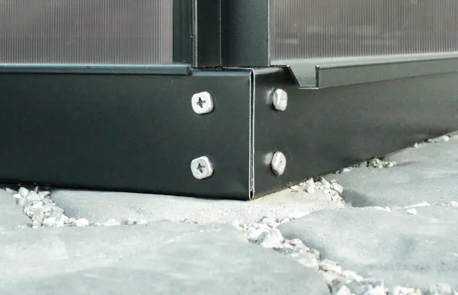 Närbild av ett metallhörnbeslag med fyra skruvar som fäster två paneler, monterat på sten- eller betongolv. Passar för konstruktion eller ramverk.