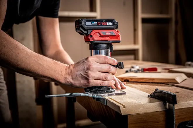 Sladdlös kompakt träfräs med ergonomiskt grepp, röd och svart design, används för exakt kantbearbetning av träskivor. Justerbart anhåll och tving för stabilitet ingår.