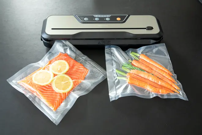 Vakuumforsegler med to poser: én med laksefileter og citronskiver, én med hele gulerødder. Perfekt til opbevaring af mad og sous vide tilberedning.