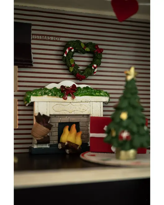 Miniature juledekoration med pejs, brænde, hjerteformet krans, indpakkede gaver og et lille juletræ med pynt og stjerne på en festlig måtte.