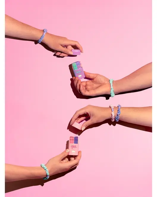 Set med pastellfärgade pilleraskar hålls av händer med flätade tygarmband mot rosa bakgrund. Varje ask har fack för daglig medicinförvaring och enkel åtkomst.