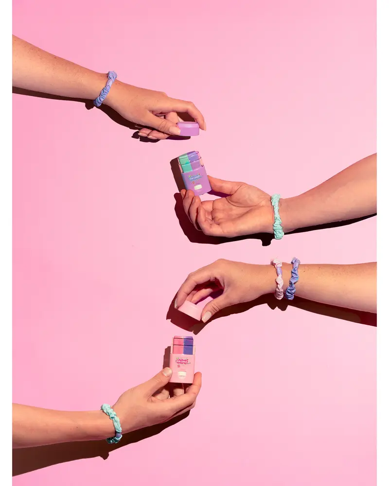 Set med pastellfärgade pilleraskar hålls av händer med flätade tygarmband mot rosa bakgrund. Varje ask har fack för daglig medicinförvaring och enkel åtkomst.