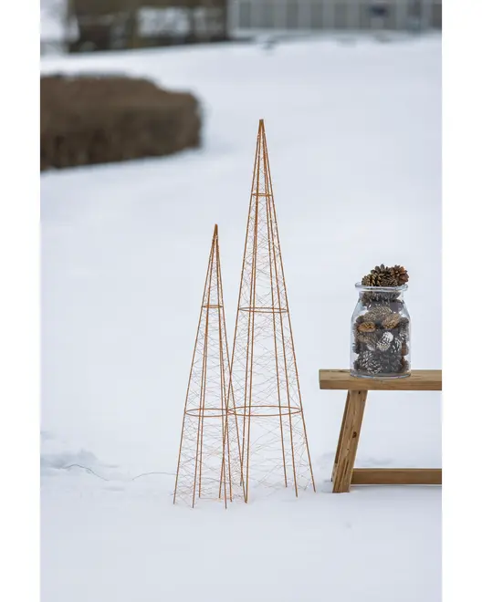 To høje, slanke plantestativer i kobberfarvet metaltråd, formet som kegler, står udendørs i sne ved siden af en træbænk med et glas fyldt med kogler.
