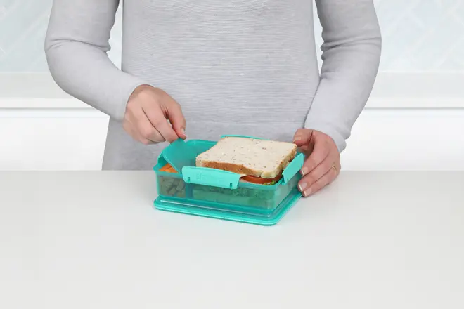 Rektangulær turkis madkasse med opdelte rum, vist med en sandwich og snacks. Sikker snaplukning. Fremstillet af holdbar plast, ideel til at holde mad adskilt.