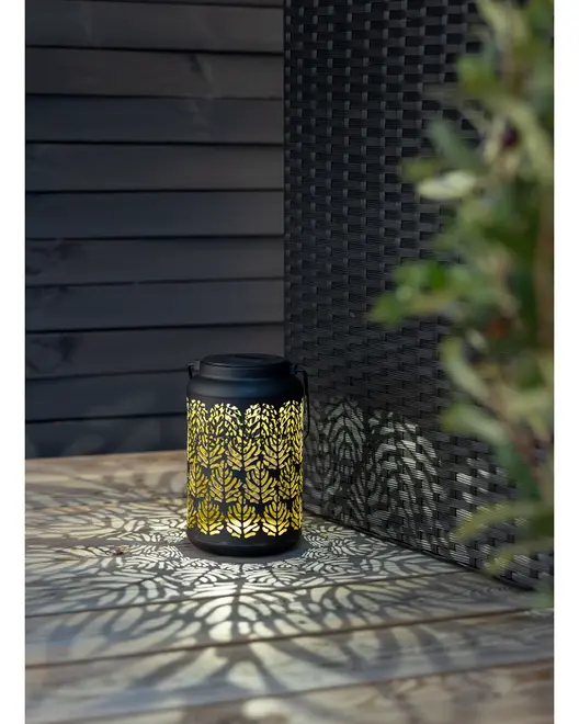 Sort metal lanterne med indviklede bladmønstrede udskæringer, udsender varmt gult lys og kaster dekorative skygger. Cylindrisk form med hank, velegnet til udendørs eller indendørs brug.