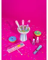 Farverigt makeup-sæt til børn med lipgloss, neglelak og pastelfarvet øjenskyggepalet, præsenteret med donut-rekvisitter og krymmel på en lyserød baggrund.