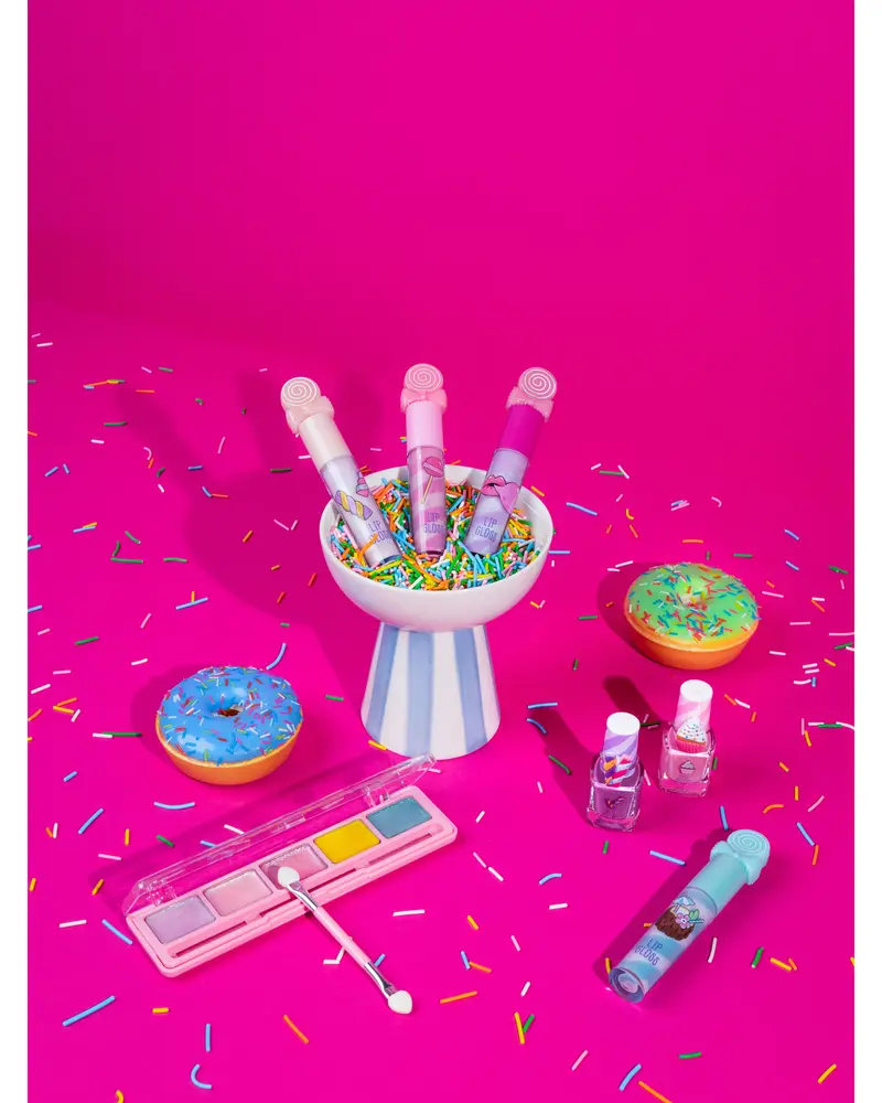 Farverigt makeup-sæt til børn med lipgloss, neglelak og pastelfarvet øjenskyggepalet, præsenteret med donut-rekvisitter og krymmel på en lyserød baggrund.