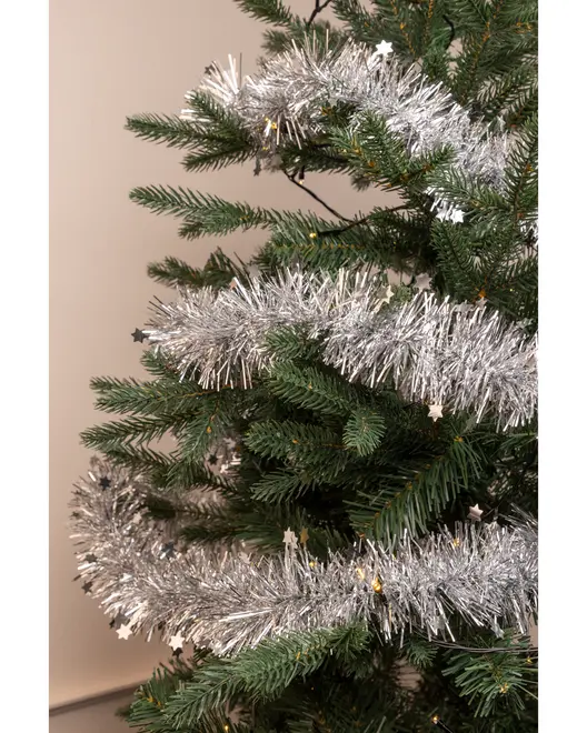 Kunstigt juletræ pyntet med tyk, sølvfarvet guirlande og små stjerneformede ornamenter. Træet har realistiske grønne grene og står op ad en lys væg.