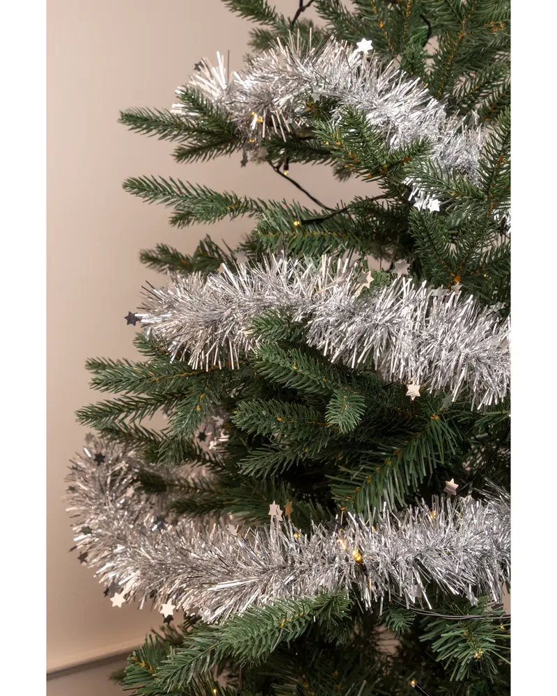 Konstgjord julgran dekorerad med tjockt, silverfärgat glitter och små stjärnformade prydnader. Granen har realistiska gröna grenar mot en ljus vägg.