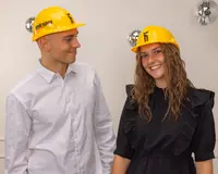 Gul byggsäkerhetshjälm i plast med justerbart band och svart tryck framtill. Blank yta, hållbar för arbete eller temafest. Ansiktet är suddigt på bilden.