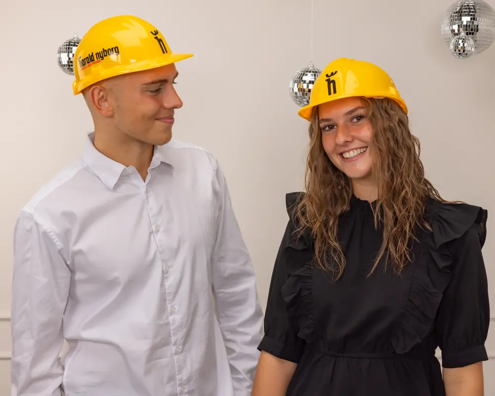 Gul byggsäkerhetshjälm i plast med justerbart band och svart tryck framtill. Blank yta, hållbar för arbete eller temafest. Ansiktet är suddigt på bilden.