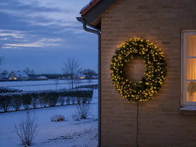 Varmhvid LED-julekrans monteret på en murstensvæg med tæt grønt løv, lysende lys og synlig ledning. - Billedet er skabt med AI
