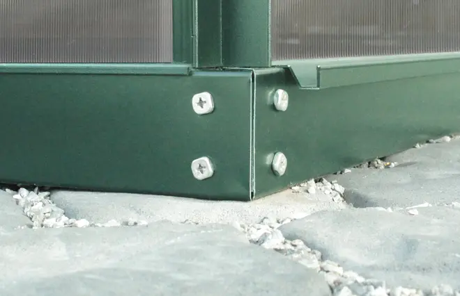 Närbild av ett grönt metallhörnbeslag med fyra skruvar som fäster två paneler. Beslaget är monterat på en sten- eller betongyta.