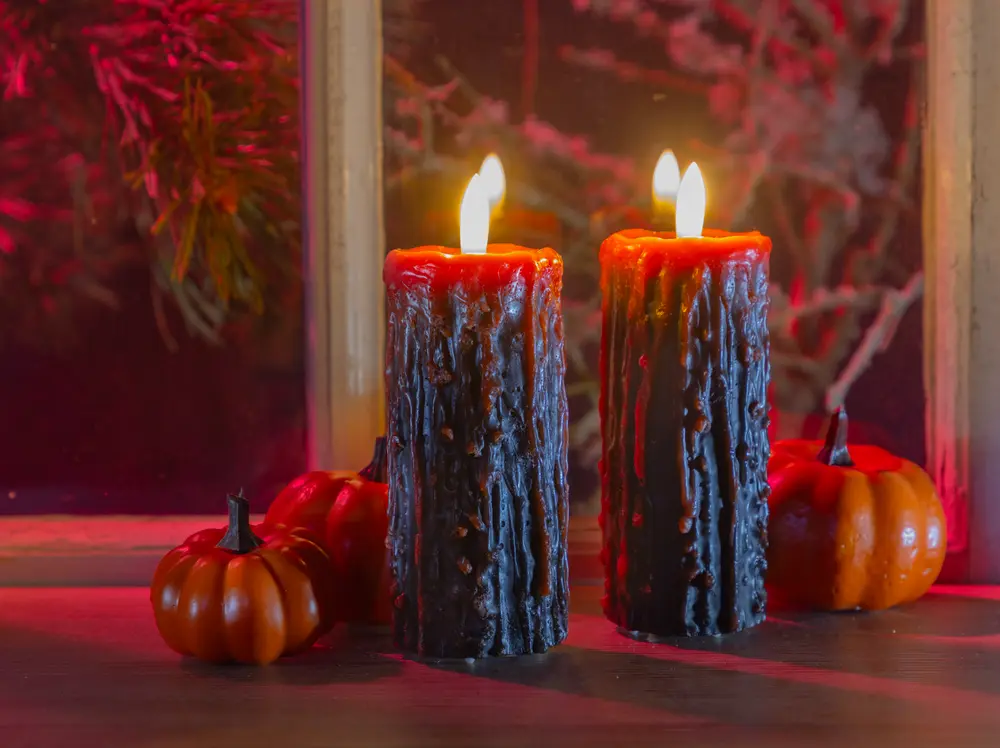 Two lit pillar candles with a textured, black wax exterior and red tops, designed to resemble tree bark. The candles have a rustic, slightly uneven surface with visible wax drips. They are displayed on a table alongside small decorative pumpkins, creating a warm, autumnal atmosphere.