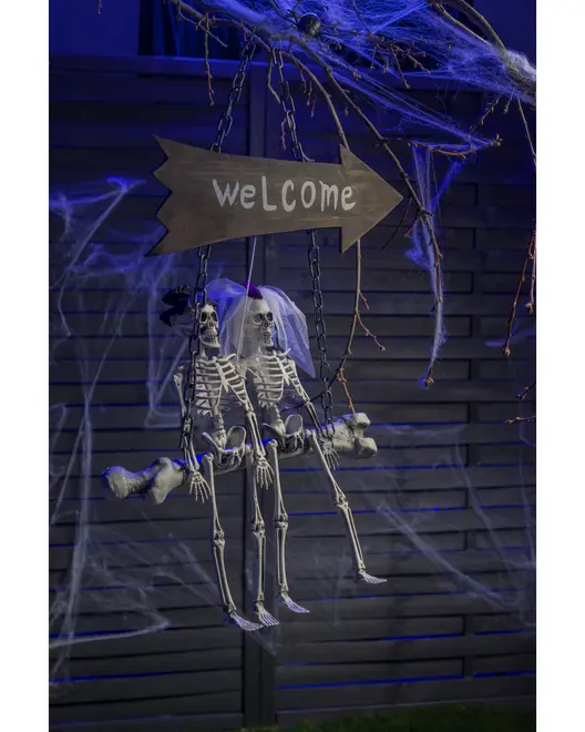 Halloween-dekoration med två skelett på en gunga, ett med slöja, under en träskylt med 'Welcome'. Omgiven av konstgjorda spindelnät, perfekt för utomhusbruk.