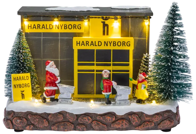 Oplyst miniature julebutik med sne, to grantræer, nissefigurer, indkøbsvogne og Harald Nyborg-skilte.