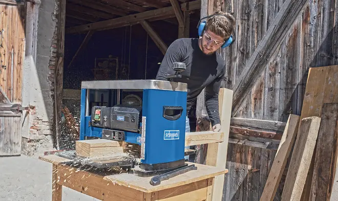 Blå elektrisk træhøvl på et træbord, bearbejder aktivt en planke. Har sidehåndtag, kontrolpanel og sikkerhedsmærkater. Velegnet til træarbejde og snedkerprojekter.