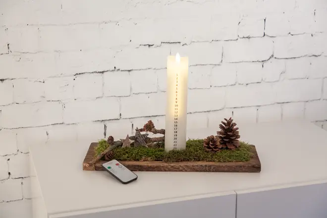 LED-stolpljus med numrerade dagar, placerat på träbricka med konstgjord mossa, kottar och dekorativa stjärnor. Fjärrkontroll ingår. Passar till juldekoration.