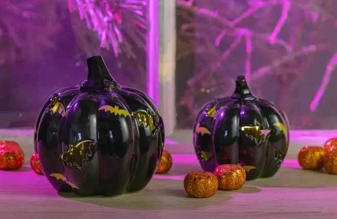 Two glossy black ceramic pumpkins with a smooth finish, decorated with shiny gold bat silhouettes. The pumpkins have a realistic ribbed texture and short, thick stems. Surrounding them are small, glittery orange decorative balls, adding a festive touch. The background features a window with purple lighting, creating a vibrant and spooky atmosphere.