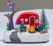 Festlig julecampingvogn med nissefigurer omkring et bål, oplyst træ, snebund og flerfarvede LED-lys.