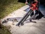 Rød SKIL rygblæser med sort opsamlingspose og langt mundstykke, vist mens den rydder blade fra en betonsti.